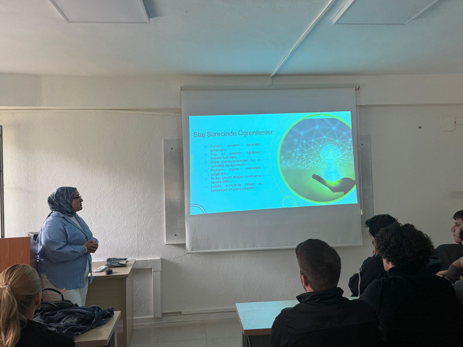 Bir öğrenci sınıf önünde durarak projeksiyonla yansıtılan slaytı anlatıyor. Slaytta ‘Staj Sürecinde Öğrenilenler’ başlıklı maddeler yer alıyor. Sınıftaki öğrenciler oturmuş, sunumu dikkatle dinliyor.