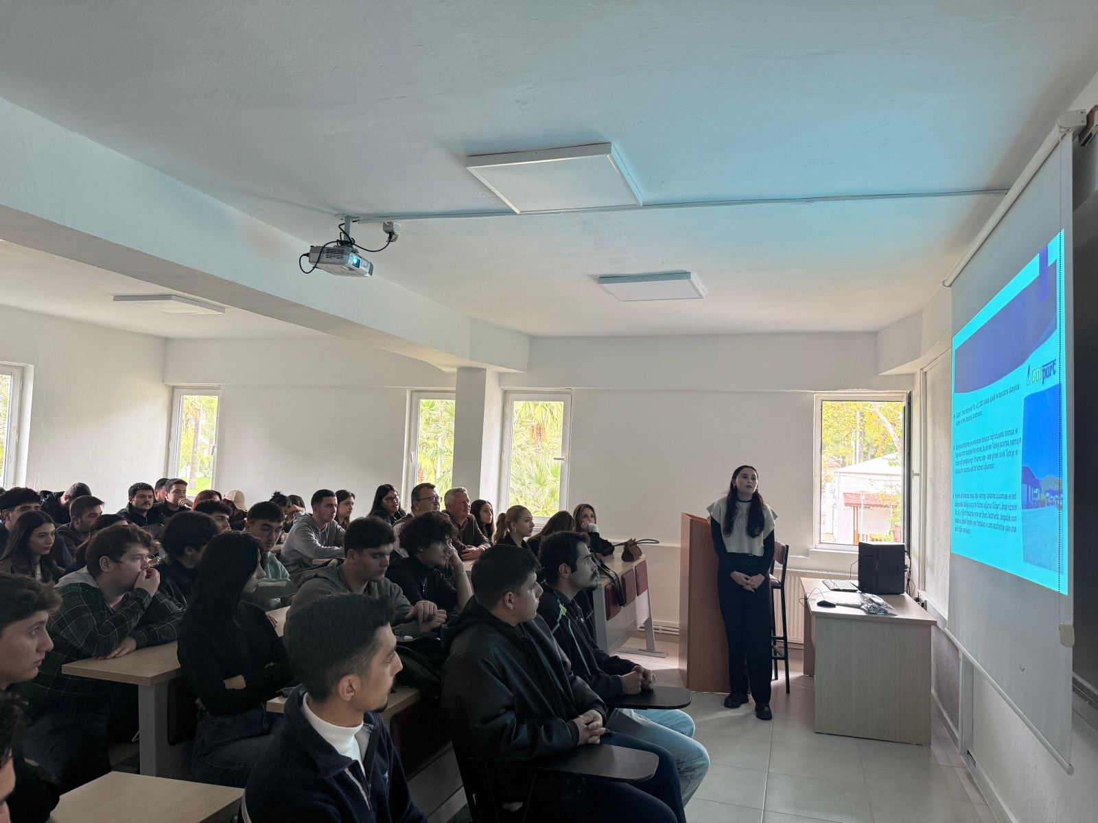 Kalabalık bir sınıfta öğrenciler sıralarda oturmuş, önde duran bir öğrencinin yaptığı sunumu izliyor. Projeksiyonda metin ve görseller bulunan bir slayt yansıtılıyor. Öğrenciler yoğun bir dikkatle sunumu takip ediyor.