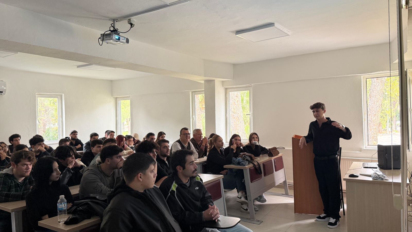 Bir öğrenci kürsü yanında ayakta durarak sunum yapıyor. Sınıf dolu ve öğrenciler sunumu dikkatle takip ediyor. Projeksiyon perdesinde sunuma ait metinler ve görseller yer alıyor.