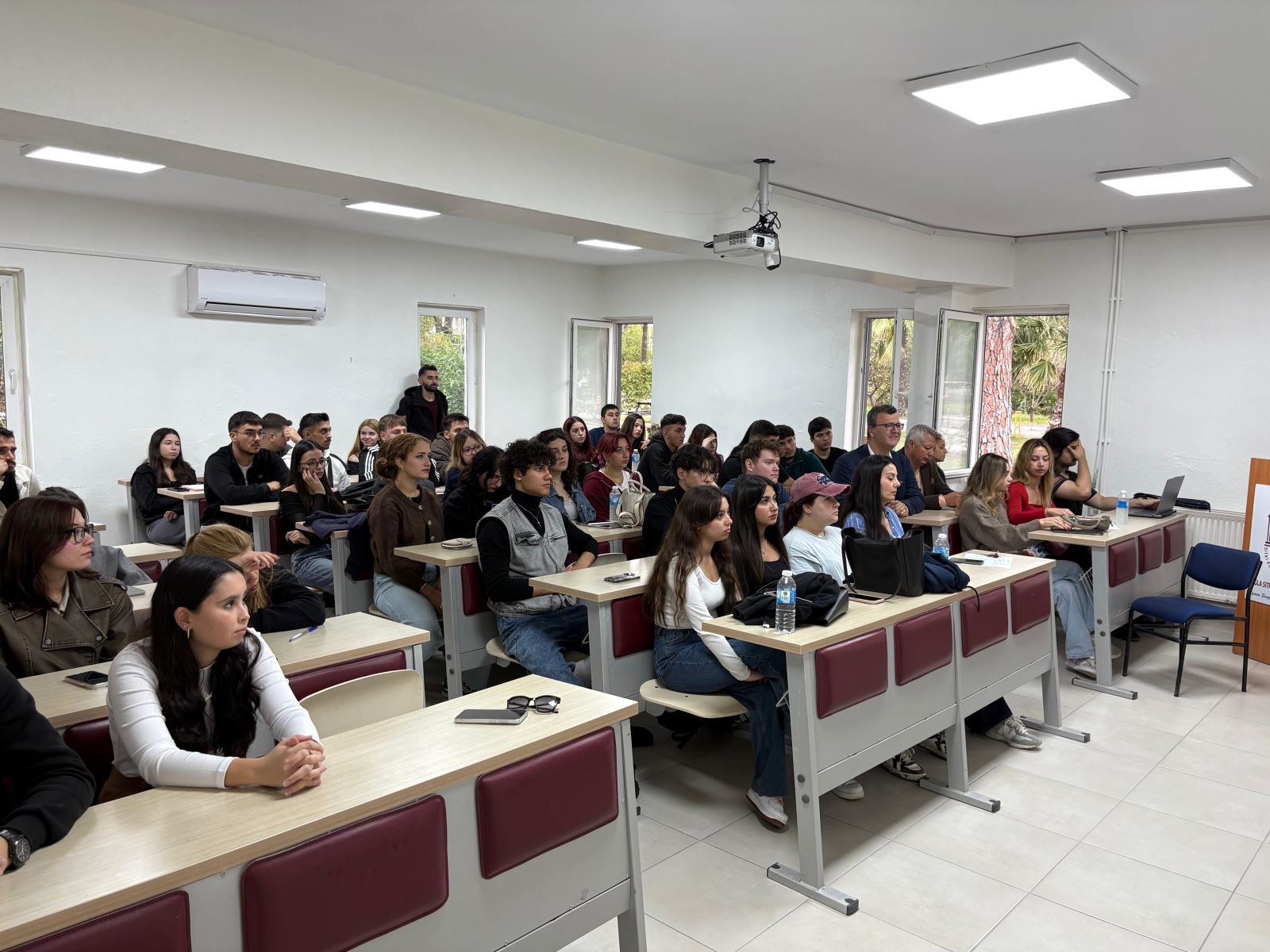 “Alumni–Student Meeting event held at Dalaman Vocational School on 23 October 2025. In a modern classroom, students are seated at desks while speaker Kudret Hasgül (Purchasing Manager at Blu Ecological Restaurant Girişim Inc.) gives a presentation. The bright classroom has white walls, a projector, and large windows allowing natural light inside.”