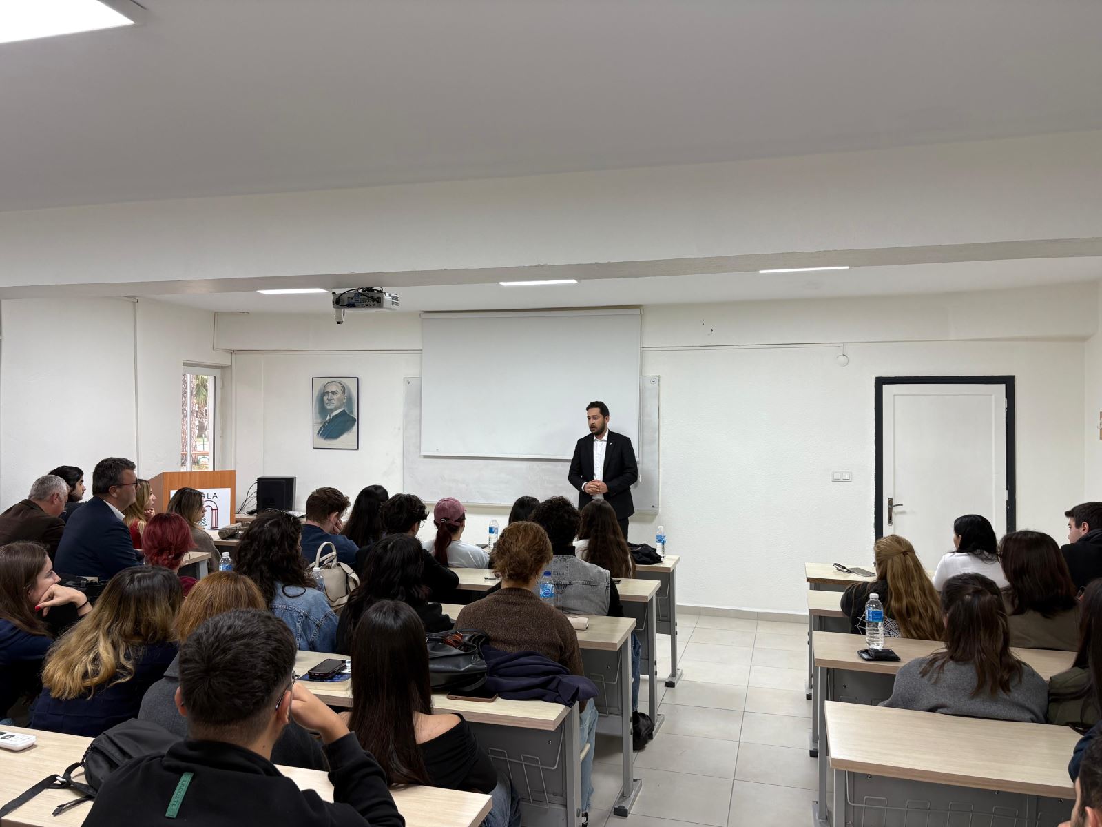 “Alumni–Student Meeting conference in a Dalaman Vocational School classroom. An Atatürk portrait and a projection screen are visible in the background. Kudret Hasgül is giving a presentation while students and faculty members listen attentively. Event dated 23 October 2025.”