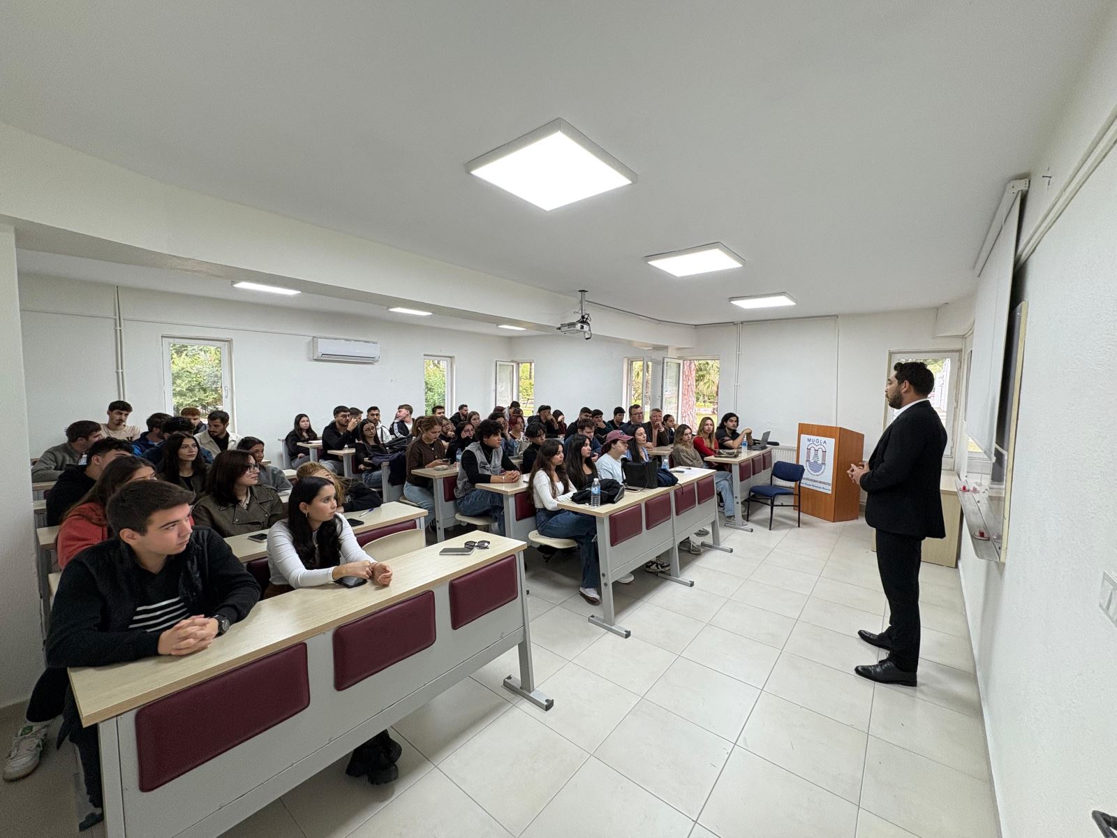 “Alumni–Student Meeting conference in a Dalaman Vocational School classroom. An Atatürk portrait and a projection screen are visible in the background. Kudret Hasgül is giving a presentation while students and faculty members listen attentively. Event dated 23 October 2025.”