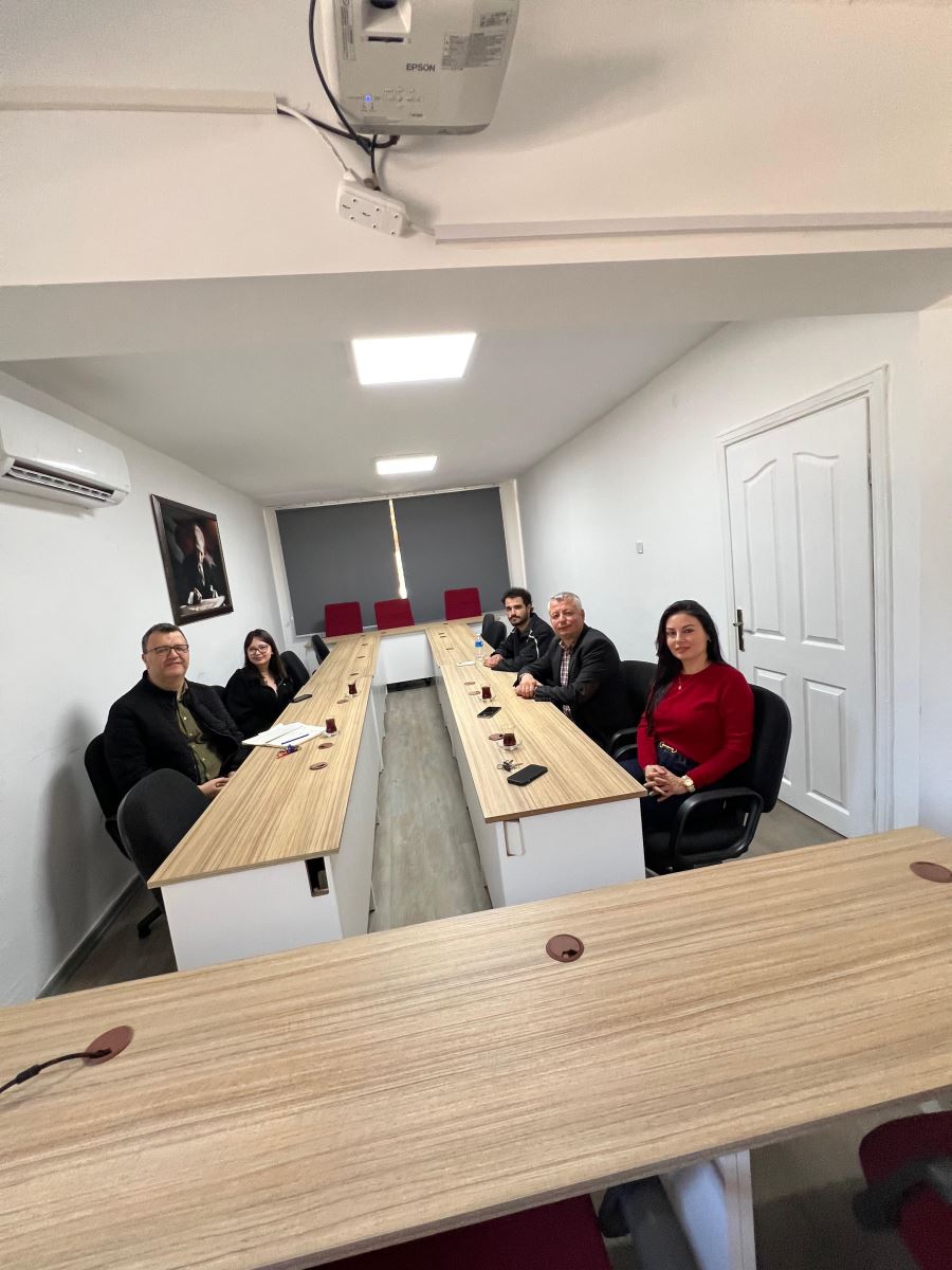 In the meeting room, five participants are seated around U-shaped tables and looking toward the camera. The bright room includes meeting tables, a wall portrait, an air conditioner, and a ceiling-mounted projector.